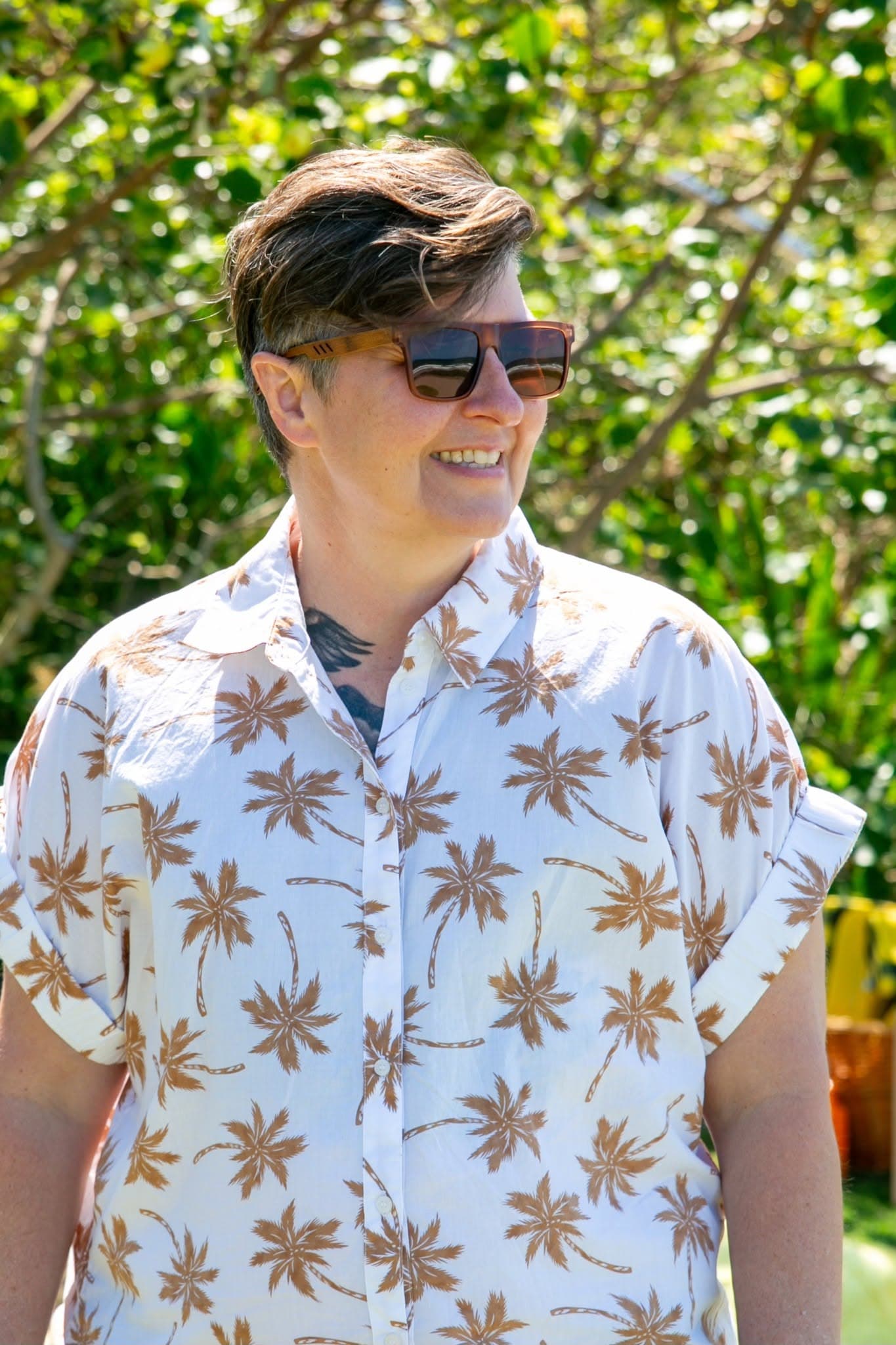 Kim Matthews outdoors, wearing sunglasses and her white shirt with a gold tropical print
